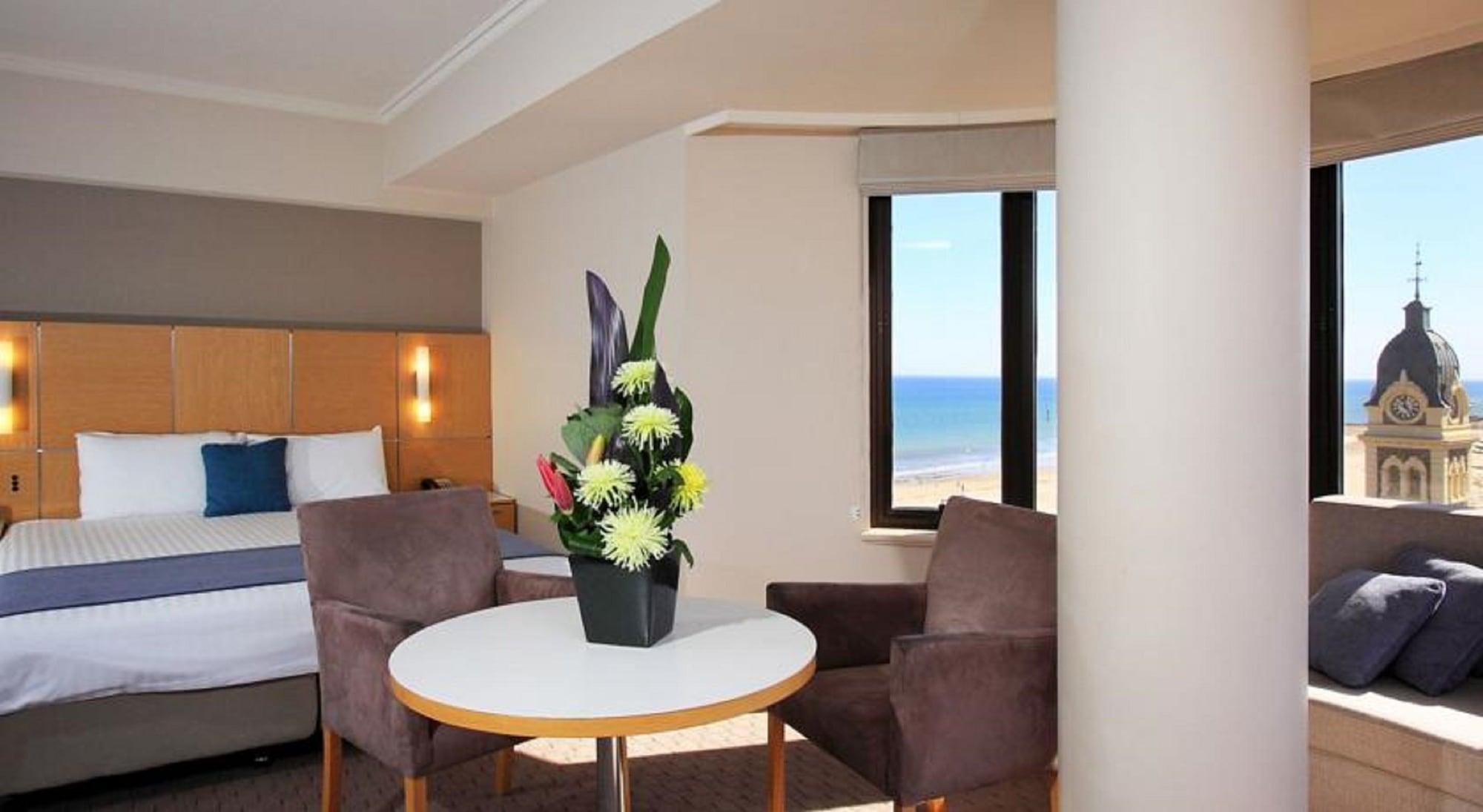 Готель Stamford Grand Adelaide Ґленелґ Екстер'єр фото The photo shows a cozy hotel room with a comfortable seating arrangement. There are two brown chairs positioned around a round table, which features a decorative flower arrangement in a black vase. The background includes a window that offers a view 