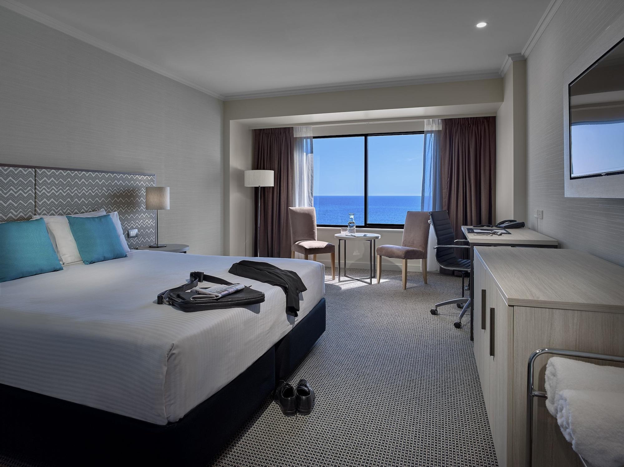 Готель Stamford Grand Adelaide Ґленелґ Екстер'єр фото The photo shows a modern hotel room featuring a queen-sized bed with white linens and two aqua blue decorative pillows. There are bedside lamps and a large window with a view of the ocean. The room also has a small seating area with two chairs and a 