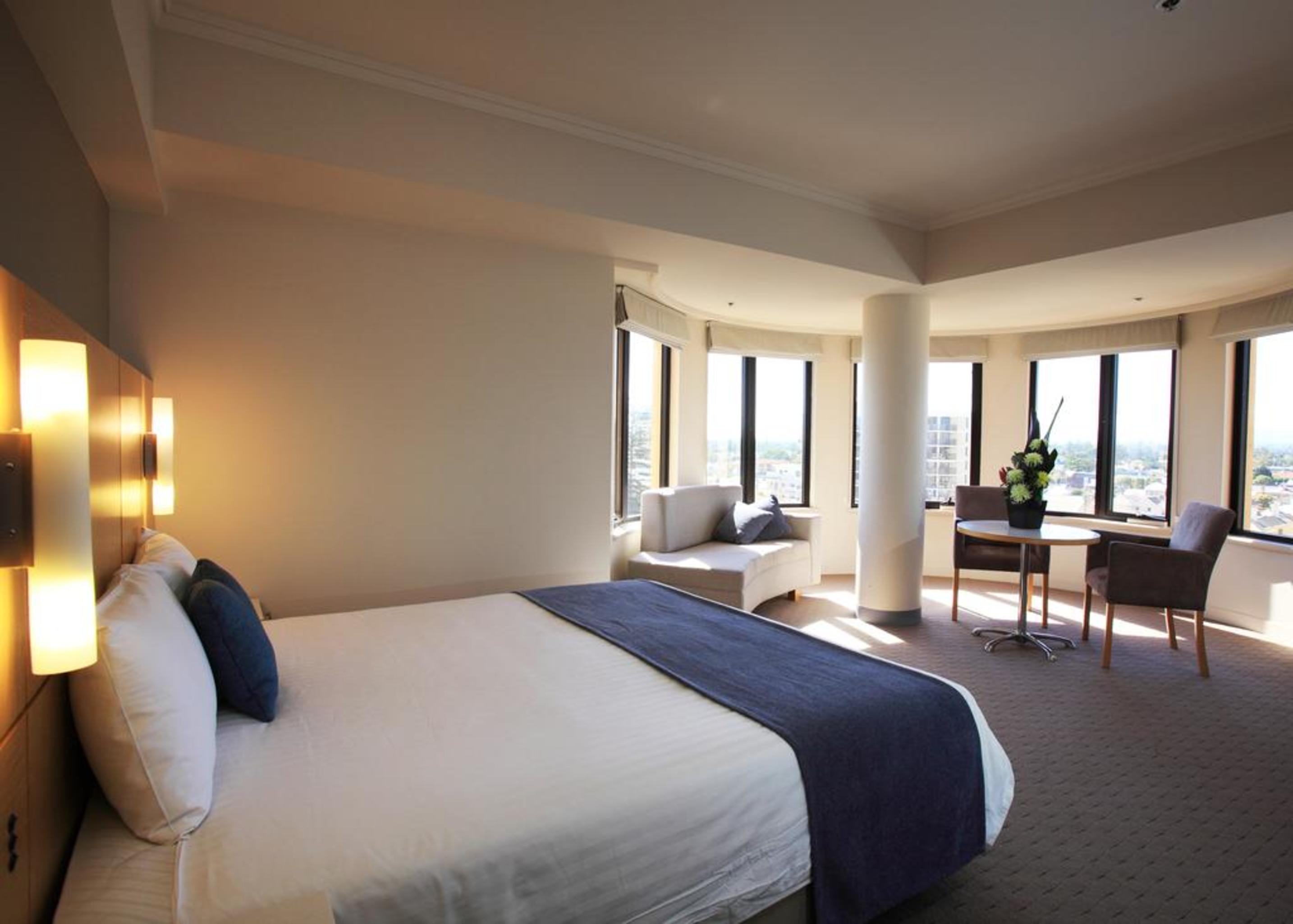 Готель Stamford Grand Adelaide Ґленелґ Екстер'єр фото The photo shows a hotel room with a modern and spacious design. There is a large bed with white linens and a dark blue throw blanket. The room features multiple windows that allow plenty of natural light, and there is a small seating area near the wi