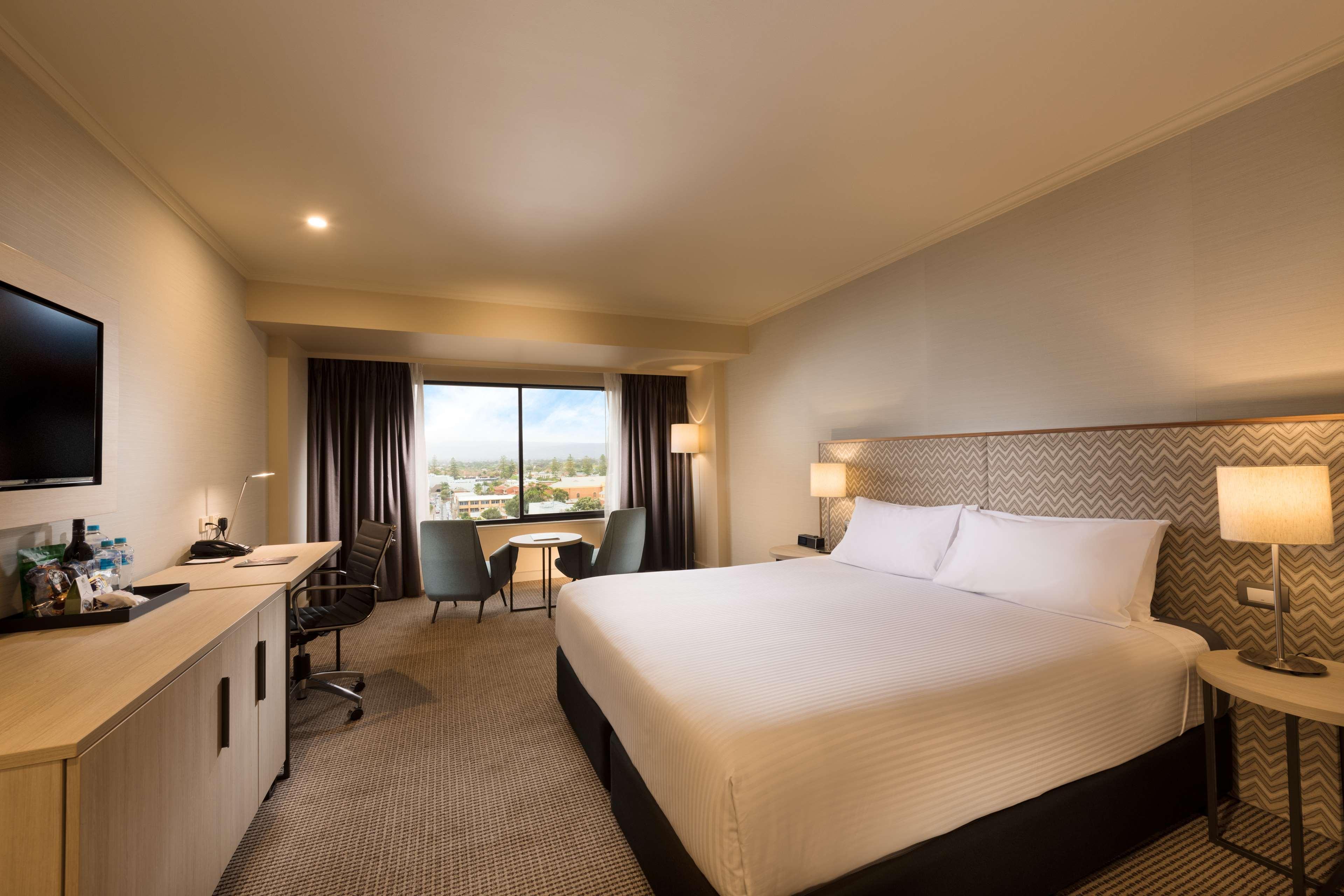 Готель Stamford Grand Adelaide Ґленелґ Екстер'єр фото The photo shows a modern hotel room. It features a large bed with white linens, a desk with a chair, and a flat-screen TV mounted on the wall. There are two armchairs and a small table near a large window that lets in natural light, providing a view 
