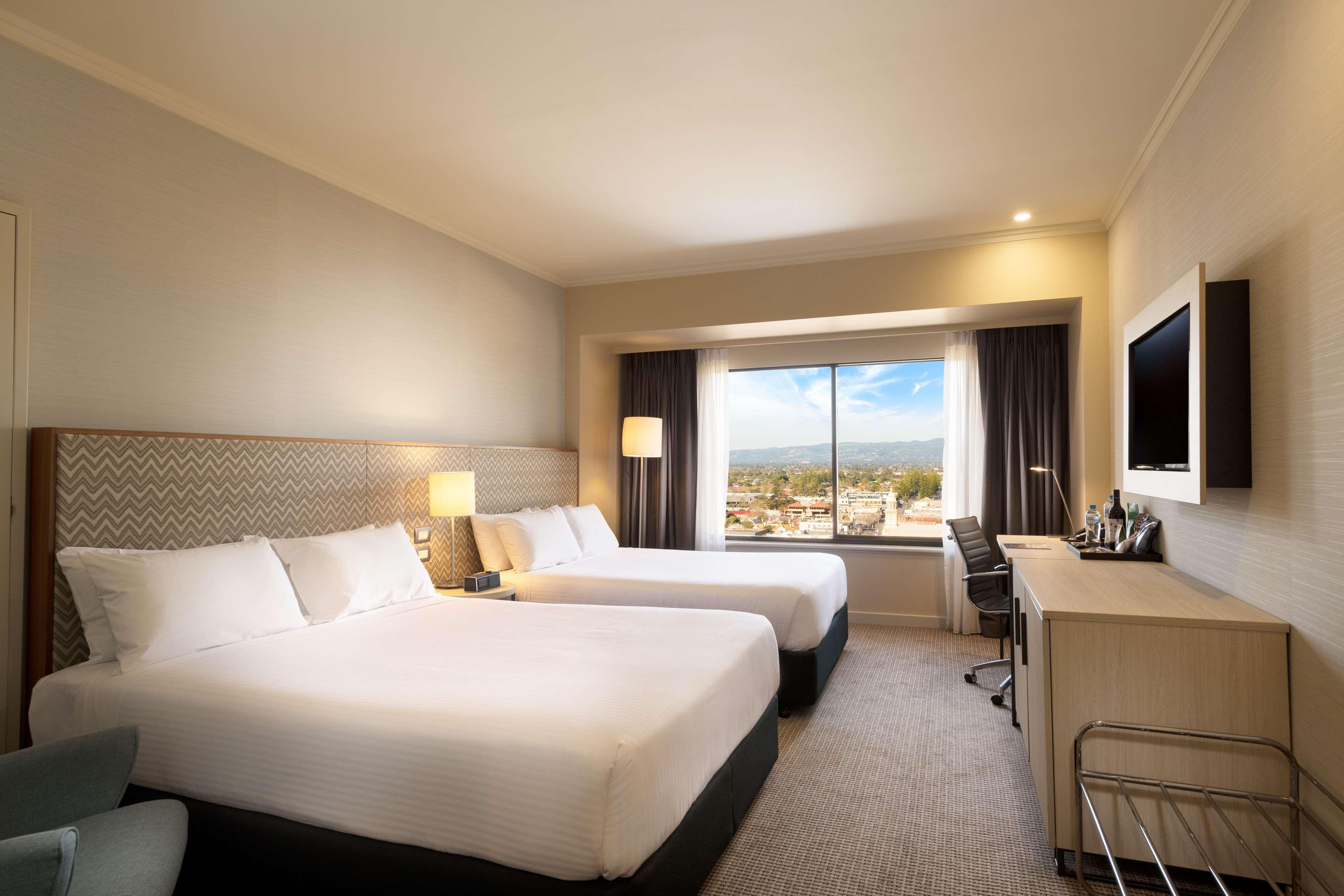 Готель Stamford Grand Adelaide Ґленелґ Екстер'єр фото The photo shows a modern hotel room featuring two beds with white linens. There are bedside tables with lamps next to each bed. A large window provides natural light and offers a view of the outside, with curtains on either side. The room has a neutr