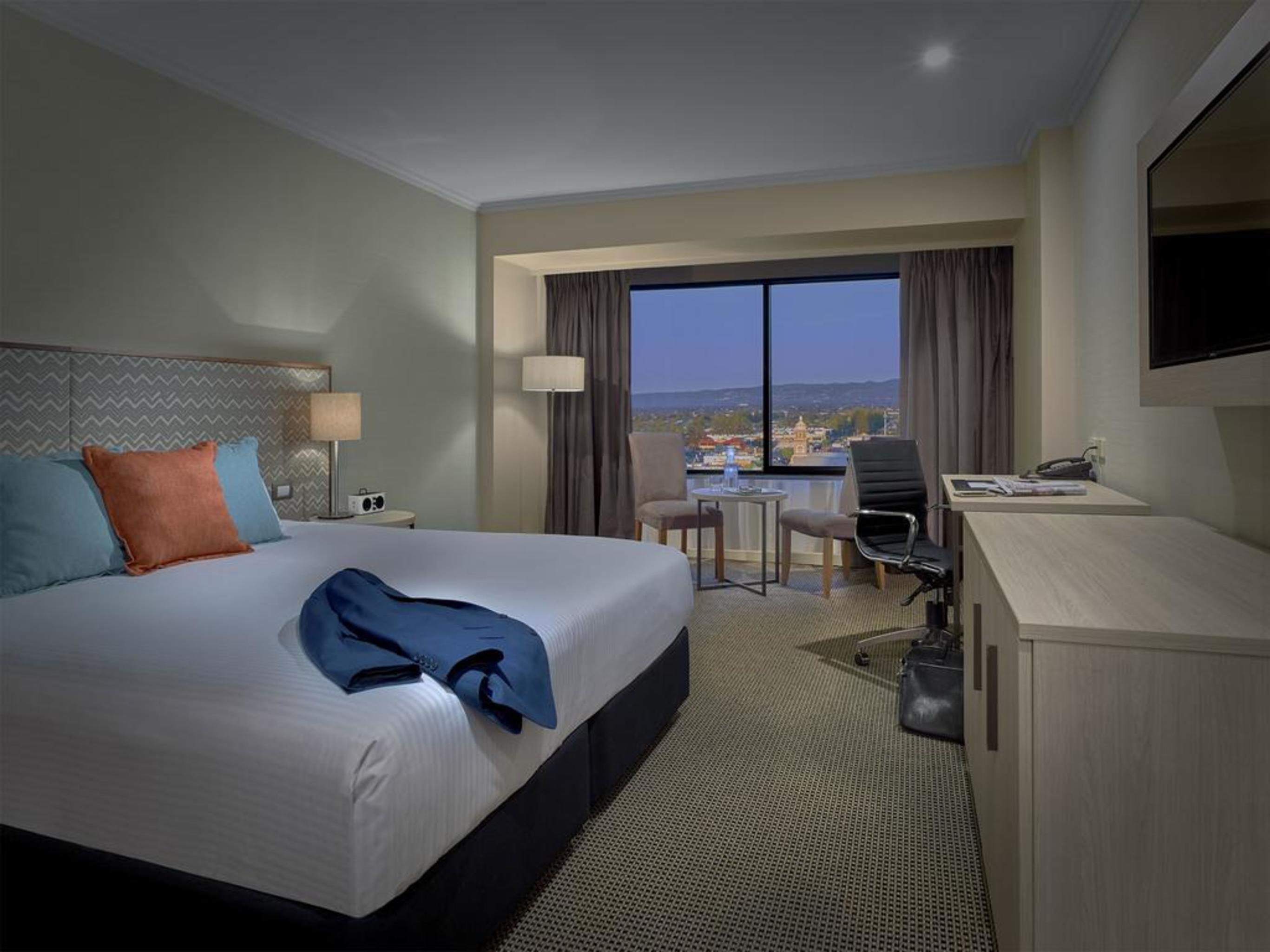 Готель Stamford Grand Adelaide Ґленелґ Екстер'єр фото The photo shows a modern hotel room. It features a large double bed with white bedding and decorative pillows in orange and blue. There's a nightstand with lamps on either side of the bed. A large window overlooks a view outside, and there's a desk a