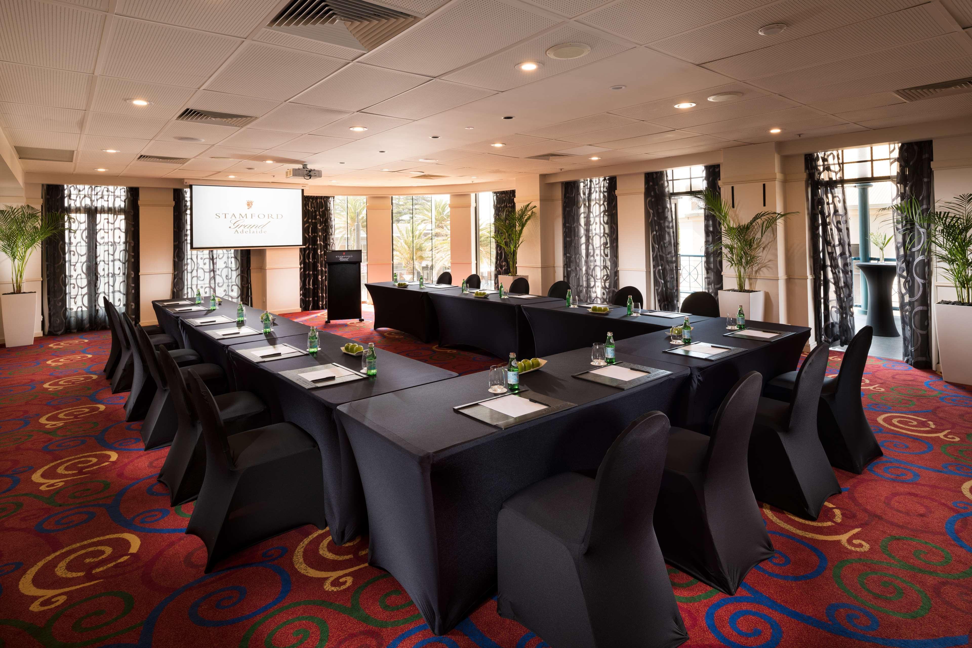 Готель Stamford Grand Adelaide Ґленелґ Екстер'єр фото The photo shows a conference room set up for a meeting or presentation. There are several tables arranged in a U-shape, covered with black tablecloths. Each table has a notepad and a small bottle of water on it. The chairs around the tables are also 