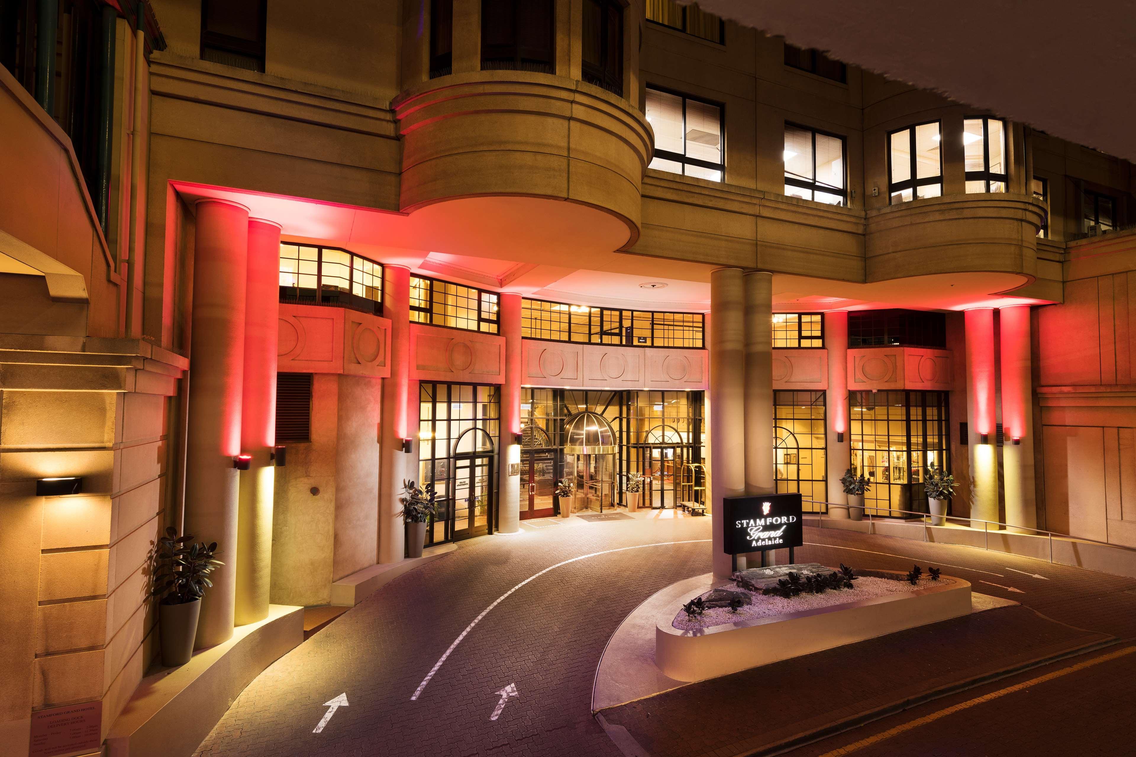 Готель Stamford Grand Adelaide Ґленелґ Екстер'єр фото The photo shows the entrance of a hotel or luxury building at night. The façade features a modern architectural style with large windows and an elegant entrance. The area is illuminated with red lighting, creating an inviting atmosphere. There appear