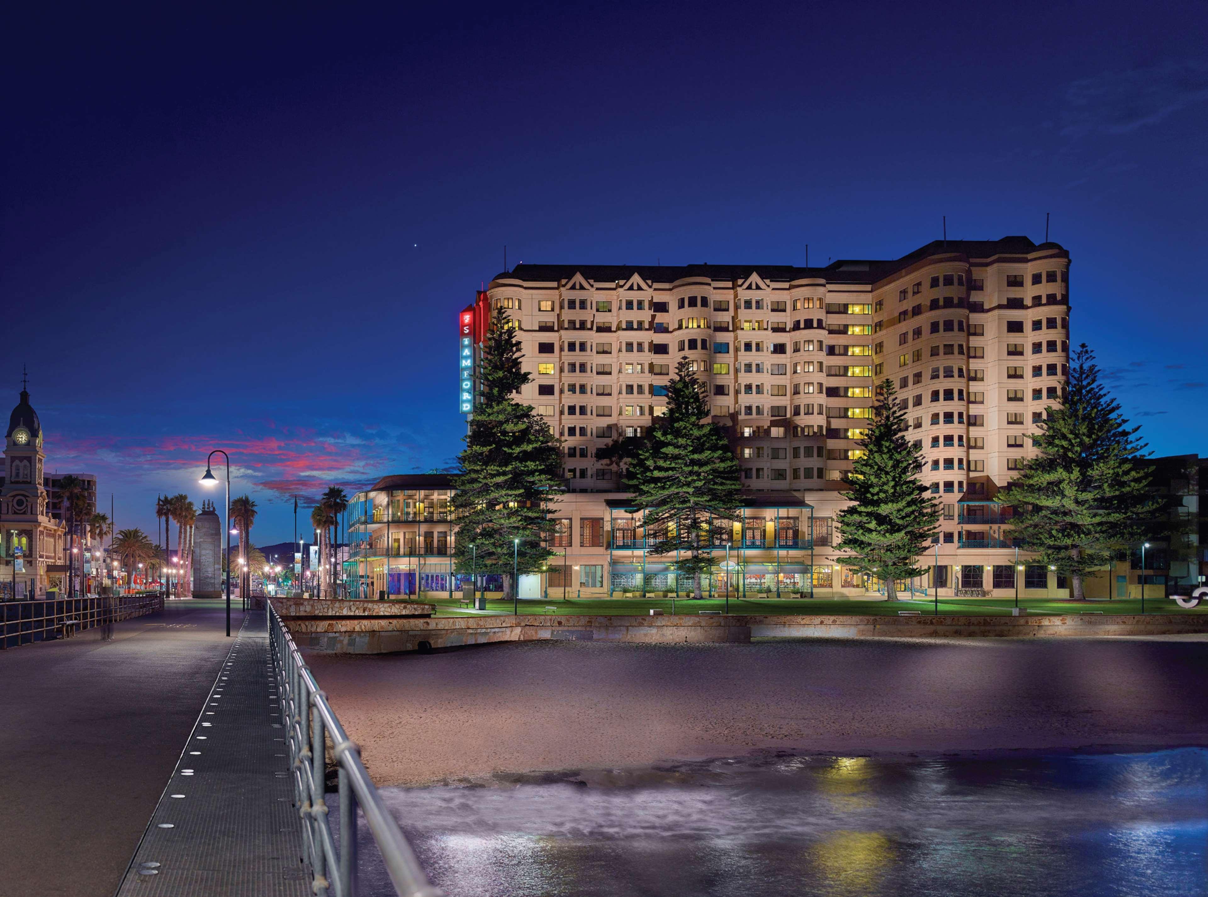 Готель Stamford Grand Adelaide Ґленелґ Екстер'єр фото The photo shows a waterfront scene at dusk, featuring a large building lit up against a darkening sky. The architecture appears modern and prominent, with several windows illuminated. In the foreground, there is a walkway or pier with fencing along t