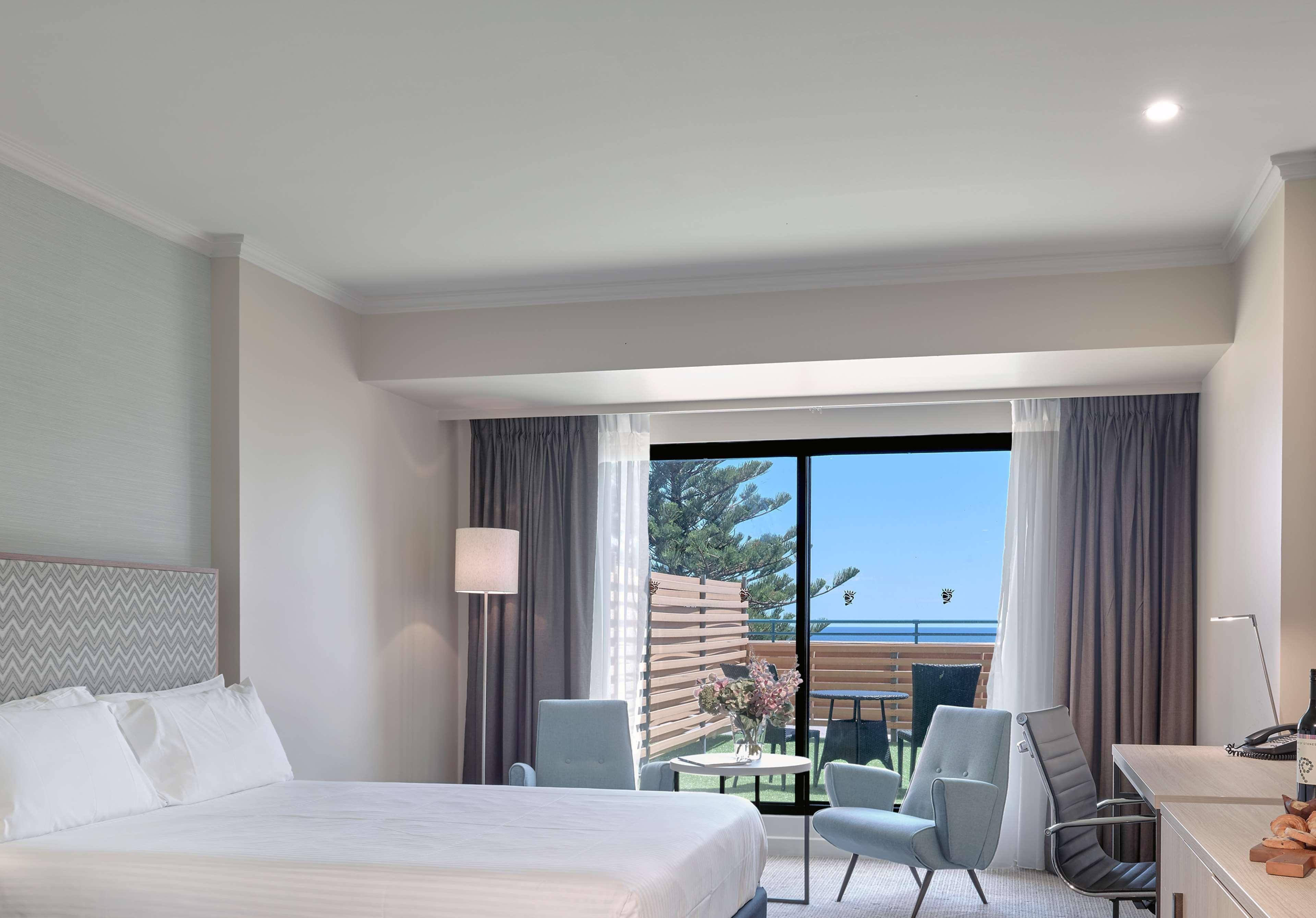 Готель Stamford Grand Adelaide Ґленелґ Екстер'єр фото The photo shows a modern hotel room featuring a large bed with white bedding. There are two armchairs positioned near a window that offers a view of an outdoor area, possibly a balcony or patio with greenery. The room has light-colored walls and curt