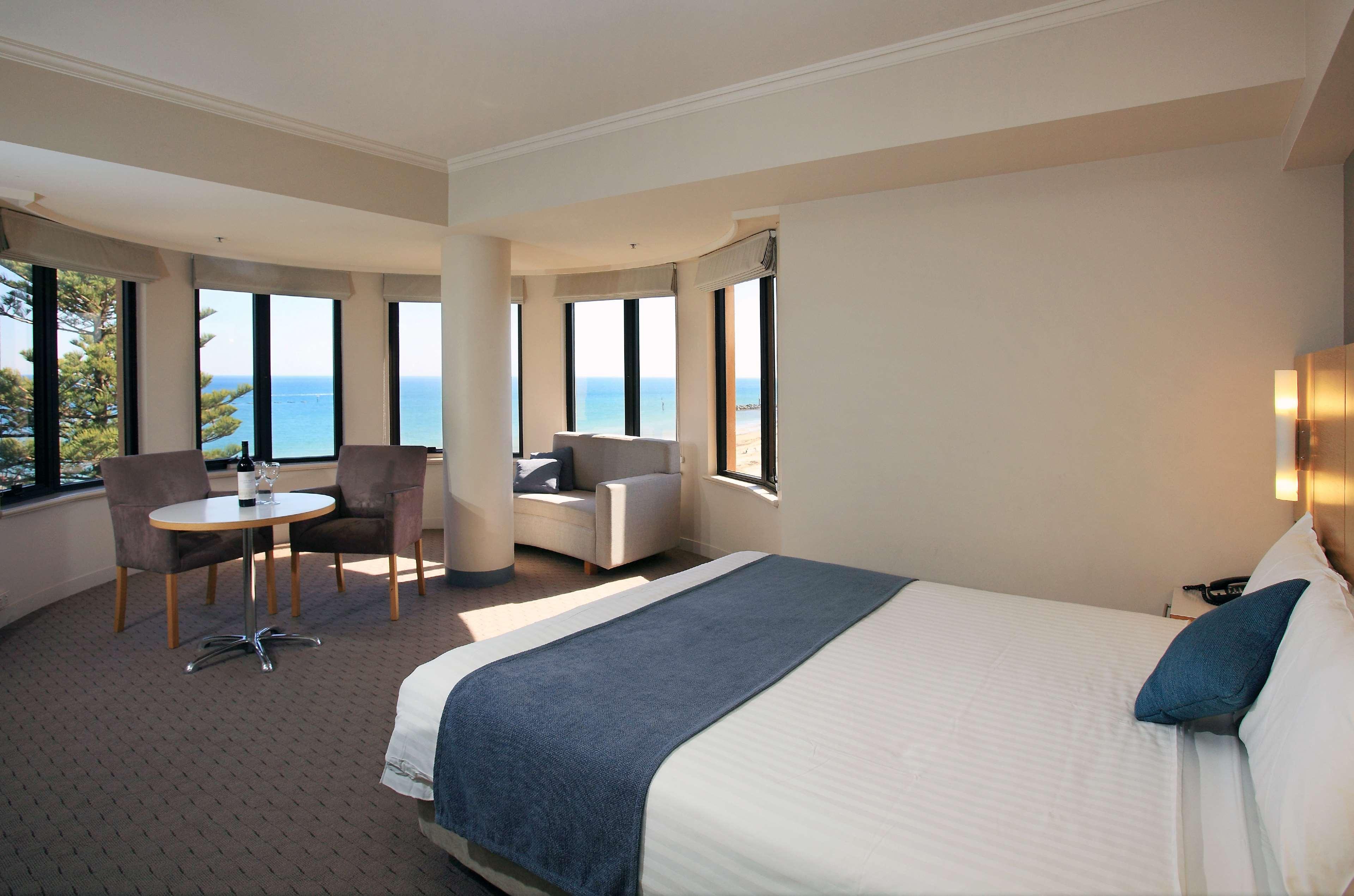 Готель Stamford Grand Adelaide Ґленелґ Екстер'єр фото The photo shows a well-lit hotel room featuring large windows that offer a view of the ocean. The room is arranged with a comfortable bed covered in white linens and a navy blue throw blanket. There is a small table and chairs set up beside the windo