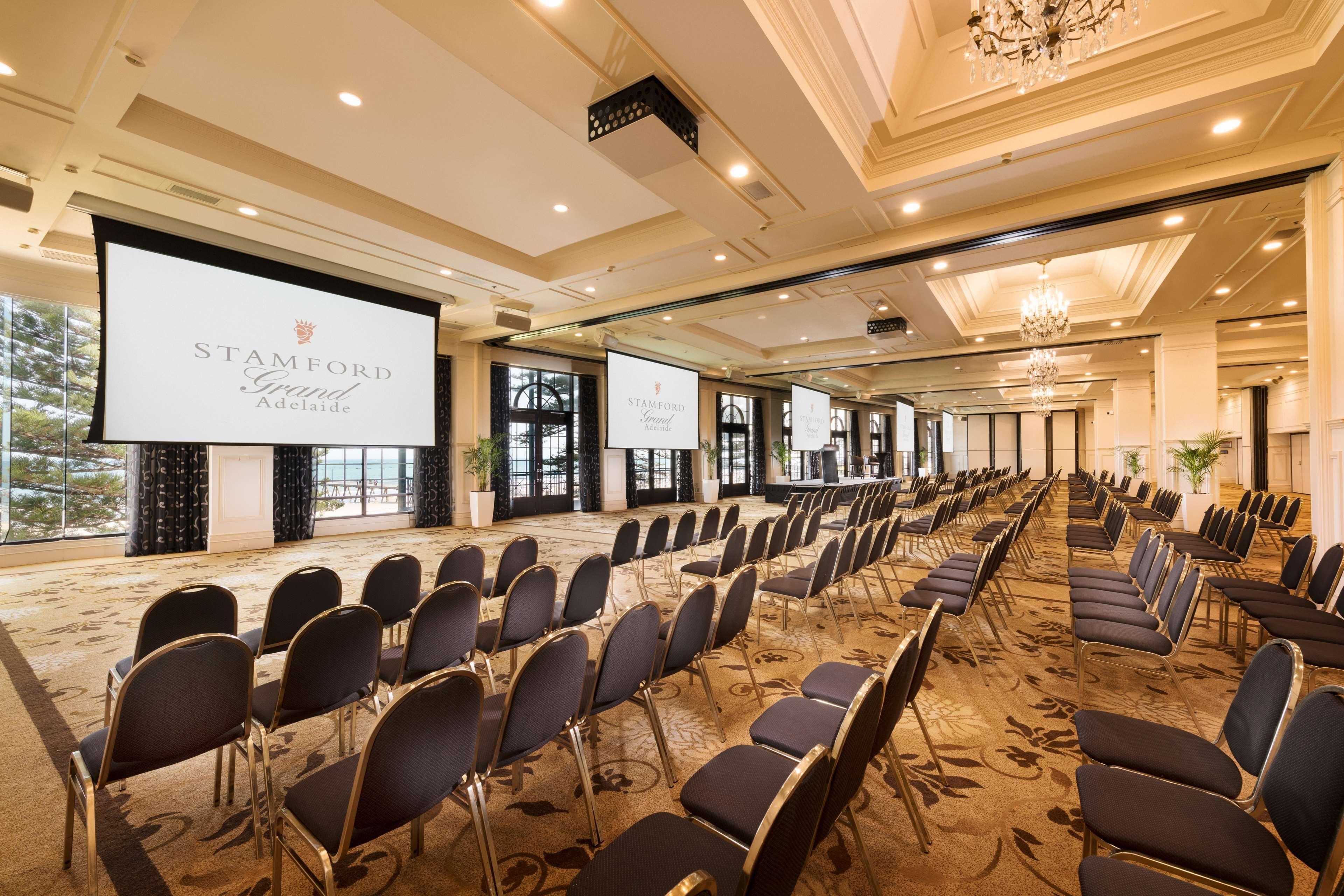 Готель Stamford Grand Adelaide Ґленелґ Екстер'єр фото The photo shows a spacious event or conference room. There are rows of dark, upholstered chairs arranged facing two large screens. The room features elegant decor with soft lighting, a chandelier, and windows that allow natural light to filter in. Th