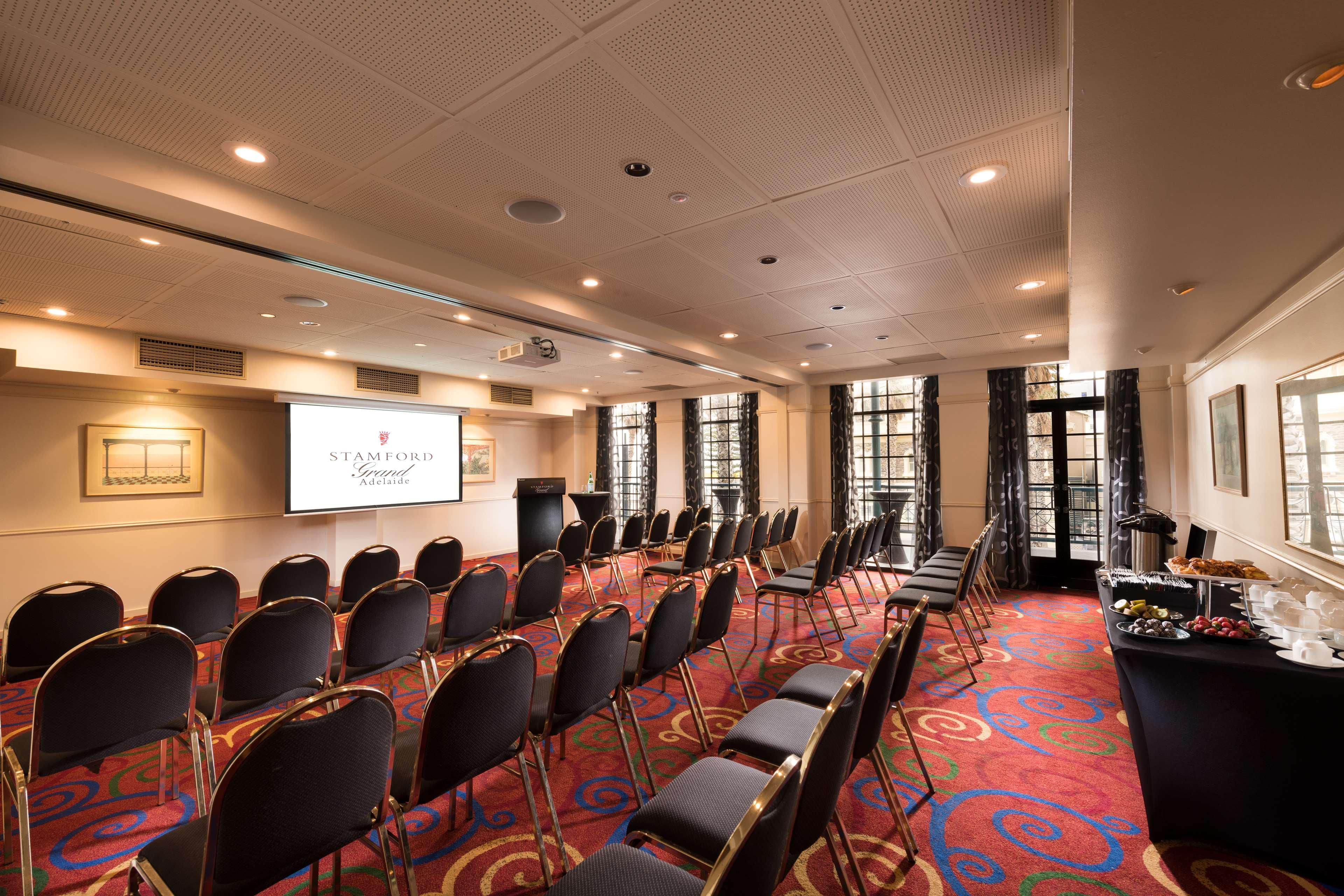 Готель Stamford Grand Adelaide Ґленелґ Екстер'єр фото The photo shows a conference room set up for an event. There are rows of black chairs arranged in a semicircular formation facing a presentation screen at the front. The room features large windows that allow natural light to enter, and the flooring 
