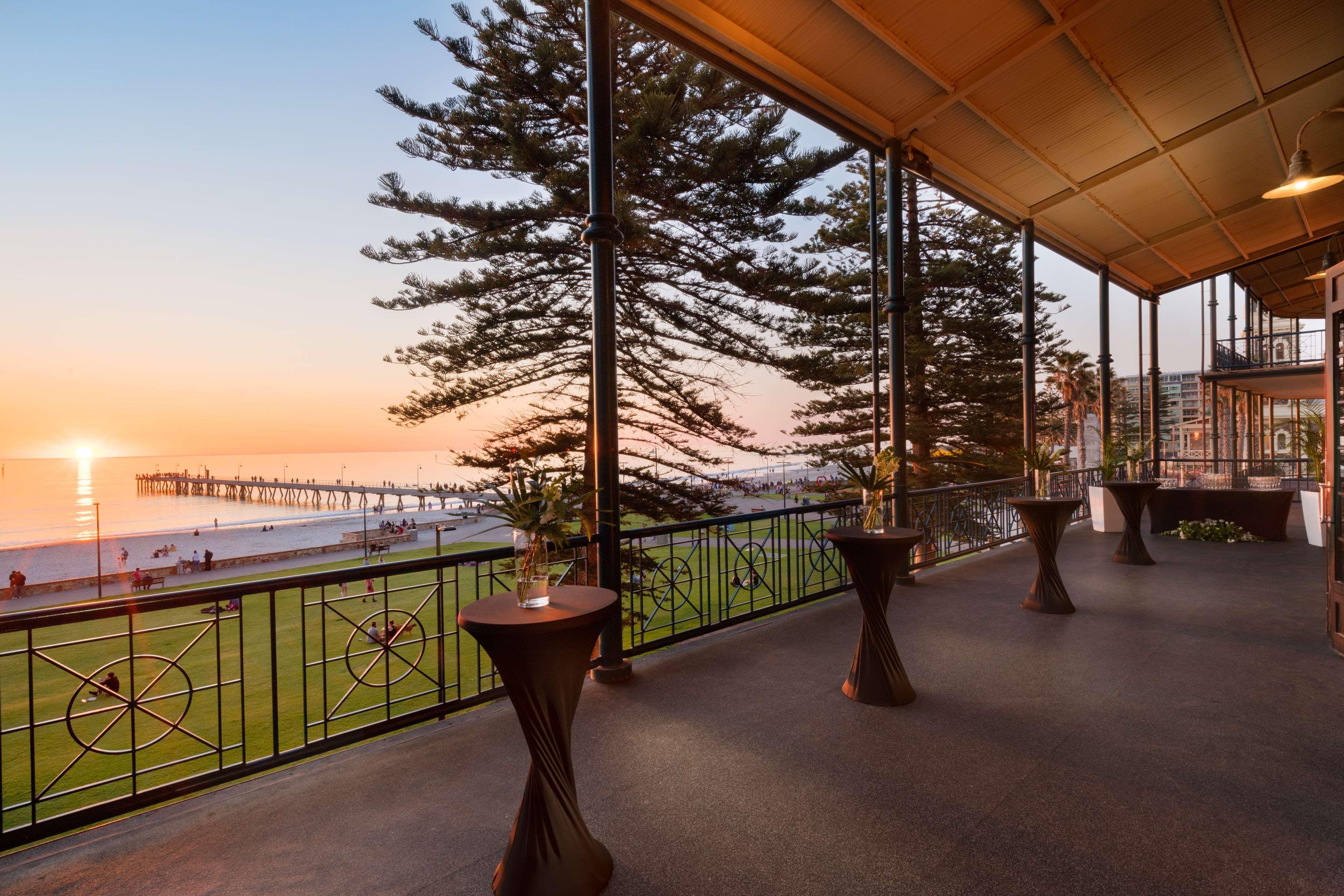 Готель Stamford Grand Adelaide Ґленелґ Екстер'єр фото The photo shows a spacious indoor space with large windows overlooking a beach at sunset. The scene features a pier extending into the ocean and a horizon painted in warm colors from the setting sun. In the foreground, there are elegant tables with d