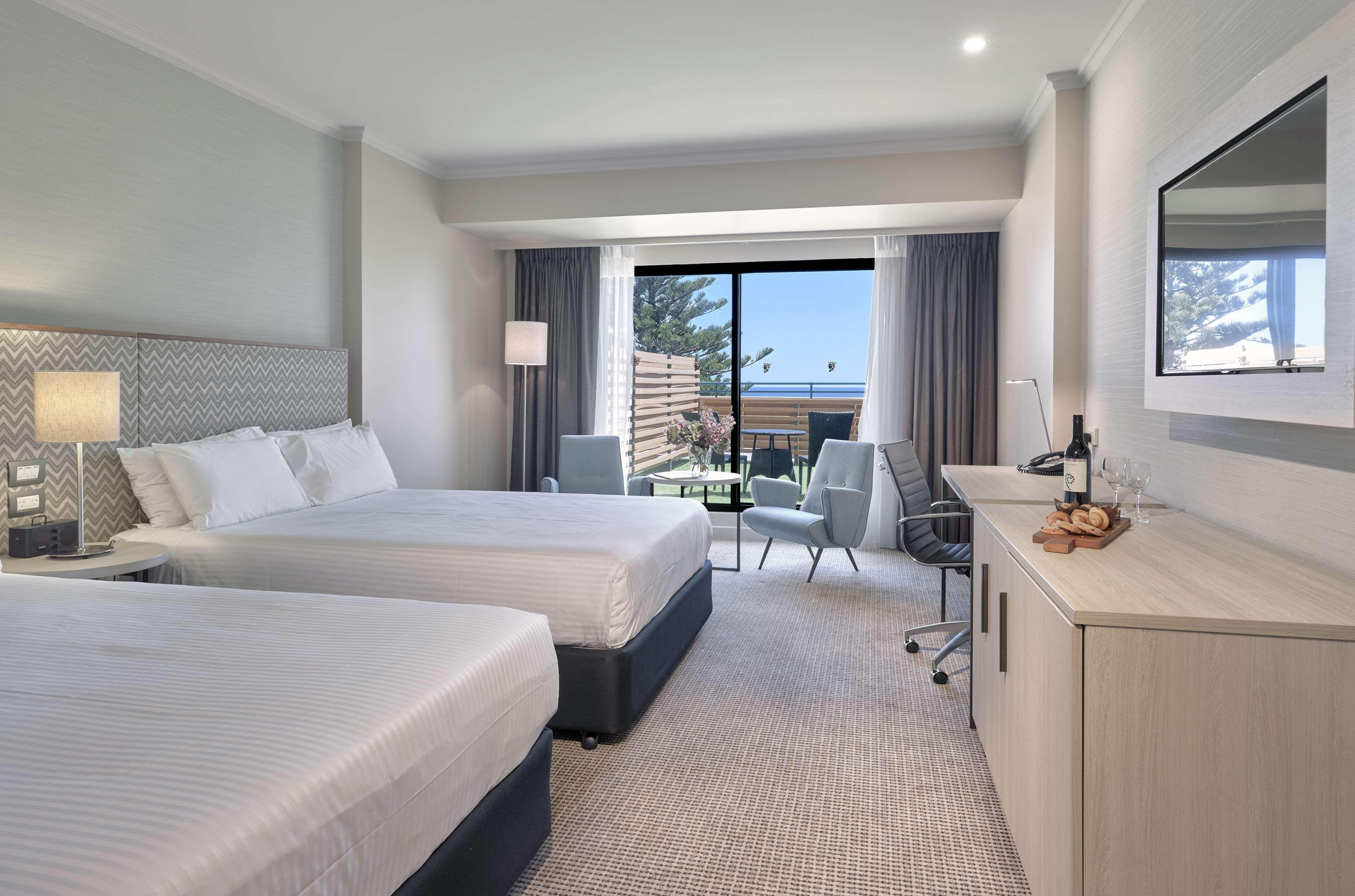 Готель Stamford Grand Adelaide Ґленелґ Екстер'єр фото The photo shows a modern hotel room featuring two beds with white linens. There's a large window that provides natural light and a view of a balcony area. The room includes two chairs and a small table, creating a cozy sitting area. A television is m