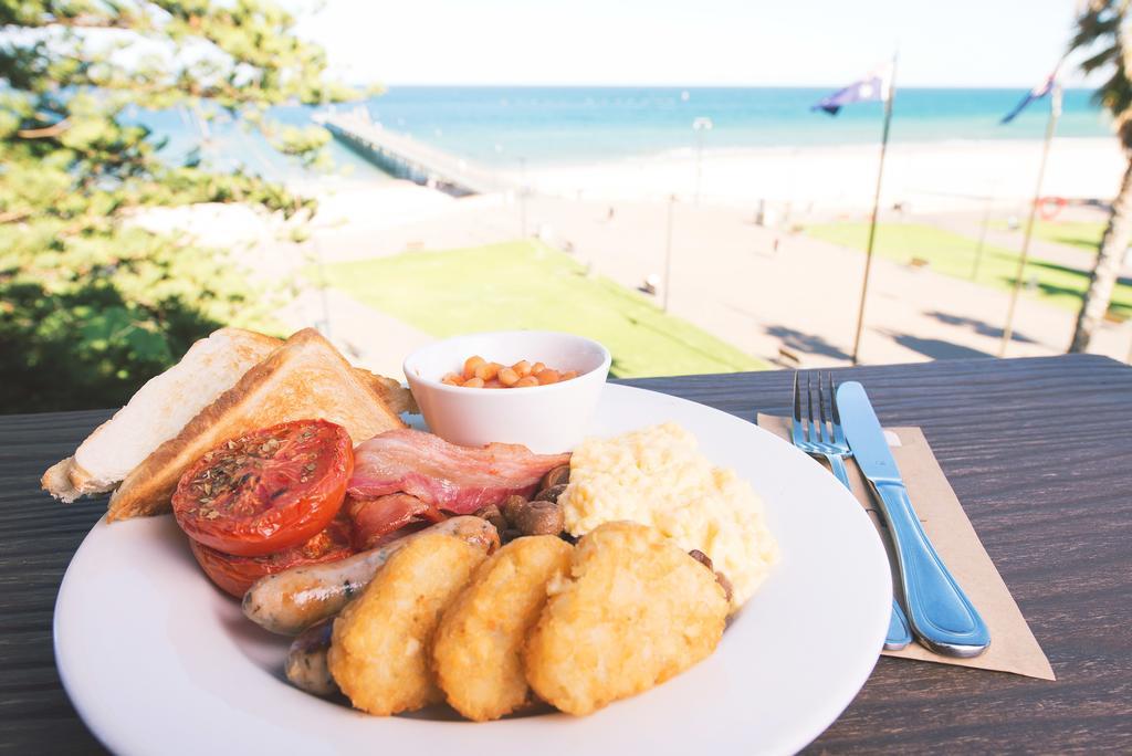 Готель Stamford Grand Adelaide Ґленелґ Екстер'єр фото The photo shows a plate of breakfast food set against a beachside backdrop. The plate includes several items: crispy bacon, a grilled tomato, sausage links, golden hash browns, and a small serving of baked beans. There is also a piece of toast on the