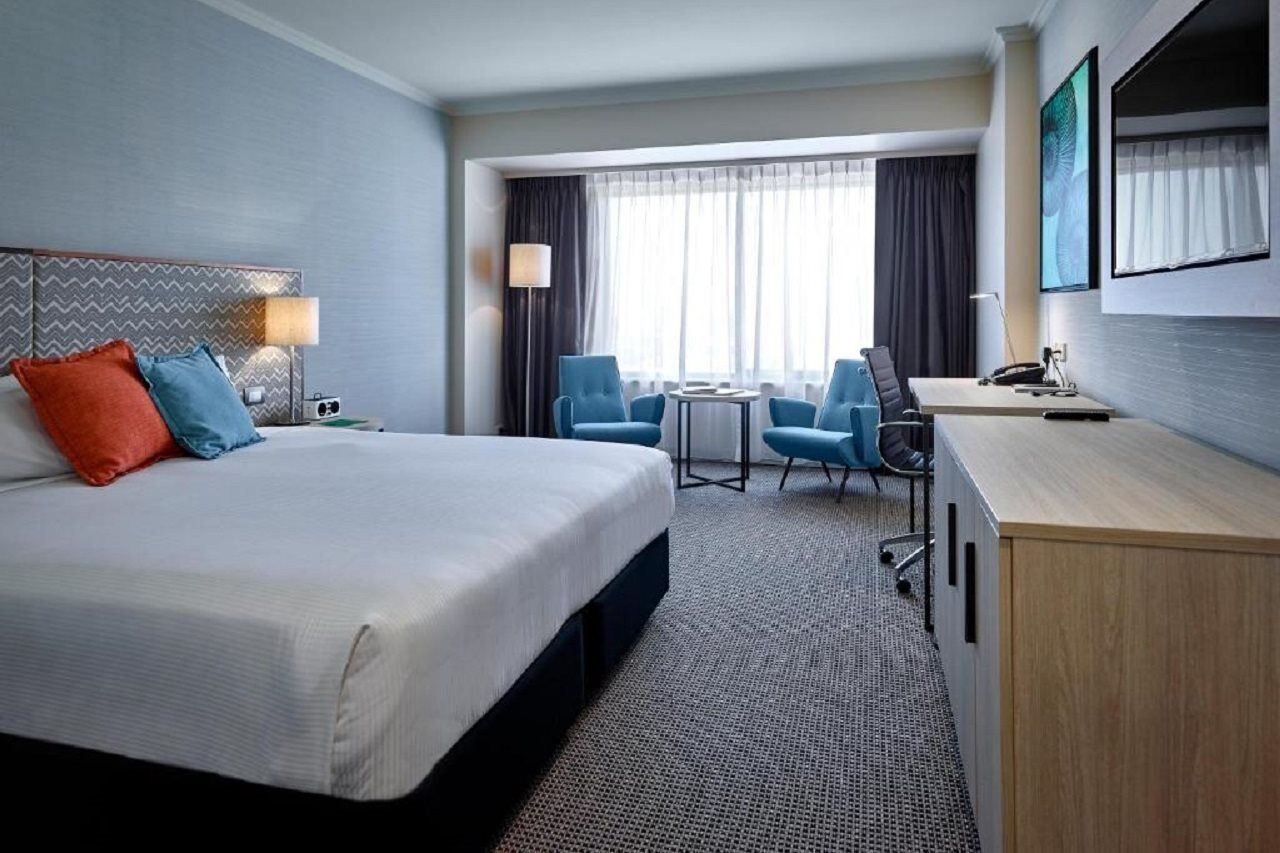 Готель Stamford Grand Adelaide Ґленелґ Екстер'єр фото The photo shows a modern hotel room with a clean and minimalist design. In the center, there is a large, neatly made bed with white linens and two decorative pillows, one orange and one blue. To the right, there is a small wooden cabinet with a telev
